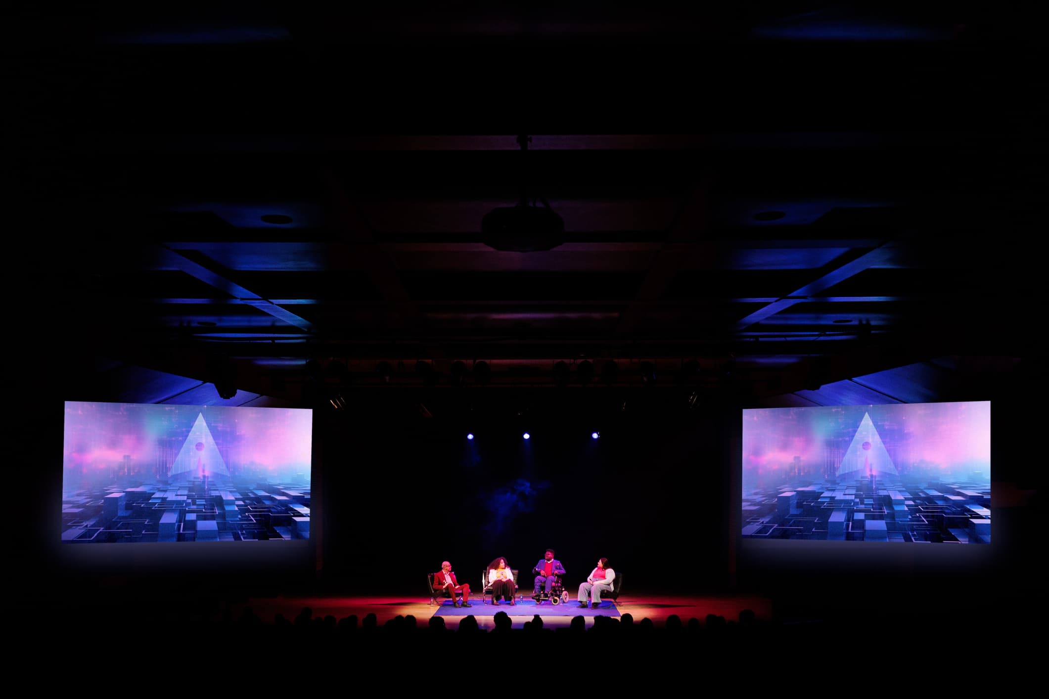 Panel de conferencia en el escenario con cuatro ponentes bajo una iluminación dramática